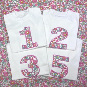 Puede incluir: Cuatro camisetas blancas de manga corta con los n&uacute;meros 1, 2, 3 y 5 bordados en grande. Cada n&uacute;mero est&aacute; relleno con un estampado floral en tonos rosa, verde y azul, delineado con un borde cosido de color rosa brillante sobre la tela.