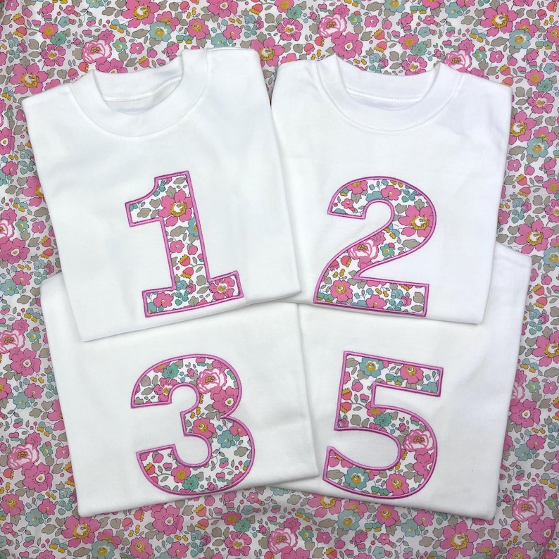 Puede incluir: Cuatro camisetas blancas de manga corta con los n&uacute;meros 1, 2, 3 y 5 bordados en grande. Cada n&uacute;mero est&aacute; relleno con un estampado floral en tonos rosa, verde y azul, delineado con un borde cosido de color rosa brillante sobre la tela.
