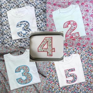 May include: A collection of white t-shirts featuring large embroidered numbers 2, 3, 4, and 5. Each number is filled with colorful floral or animal-themed fabric patterns and outlined with decorative stitching, displayed on various patterned fabrics.