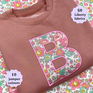 May include: A dusty rose sweatshirt featuring a large letter B appliqued in a floral Liberty fabric with pink, green, and yellow blossoms. The letter is outlined in bright pink stitching. Text on the image reads 50 Liberty fabrics and 10 jumper colours.