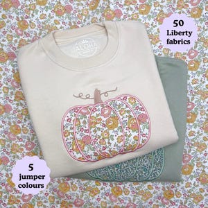 Peut inclure: Deux sweat-shirts pliés, crème et vert sauge, sont empilés. Chacun présente une citrouille brodée avec du tissu floral. Le sweat-shirt crème porte le texte "ALOHA" sur le col. L'image affiche également le texte "5 couleurs de pull" et "50 tissus Liberty".