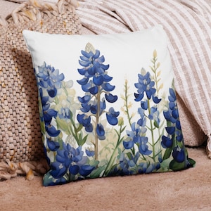 May include: A white throw pillow with a blue and green floral design featuring bluebonnet flowers. The pillow is on a tan couch with a white and tan striped throw blanket.
