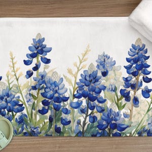 May include: A white bath mat featuring a watercolor design of bluebonnet flowers. The floral pattern includes blue and white flowers with green stems and leaves. A white towel and a pair of light green sandals are also visible.