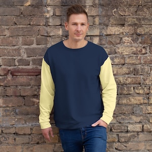 May include: A navy blue sweatshirt with pale yellow sleeves. The person in the image is wearing blue jeans. The sweatshirt has a crew neck and a relaxed fit. The background is a brick wall.