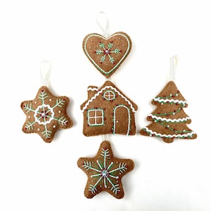 May include: Set of five felt gingerbread ornaments, including a heart, a house, a star, a tree, and a snowflake. Each ornament is decorated with white icing, green sprinkles, and red beads.