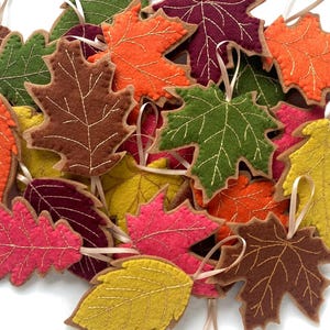 Peut inclure: Une collection d'ornements en feutrine en forme de feuilles aux couleurs automnales variées, notamment orange, jaune, vert, marron, rose et rouge foncé. Chaque feuille présente des coutures dorées décoratives et une petite boucle en ruban.