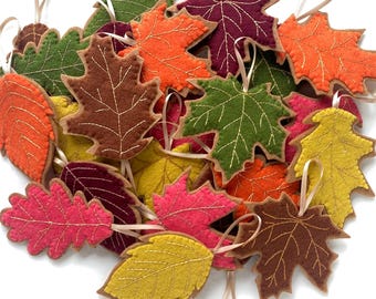 May include: A collection of felt leaf ornaments in various shapes and autumn colors including orange, yellow, green, brown, pink, and deep red. Each leaf features decorative gold-colored stitching along the veins and a small ribbon loop for hanging.