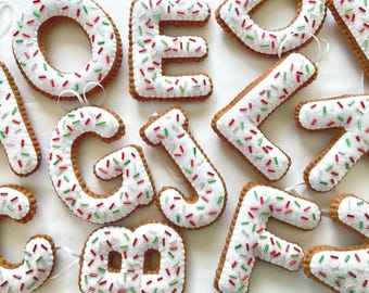 May include: A collection of felt alphabet letters shaped like cookies with white icing and red and green sprinkles. Each letter features a brown base, a white top layer, and small attached white ribbons for hanging as ornaments.
