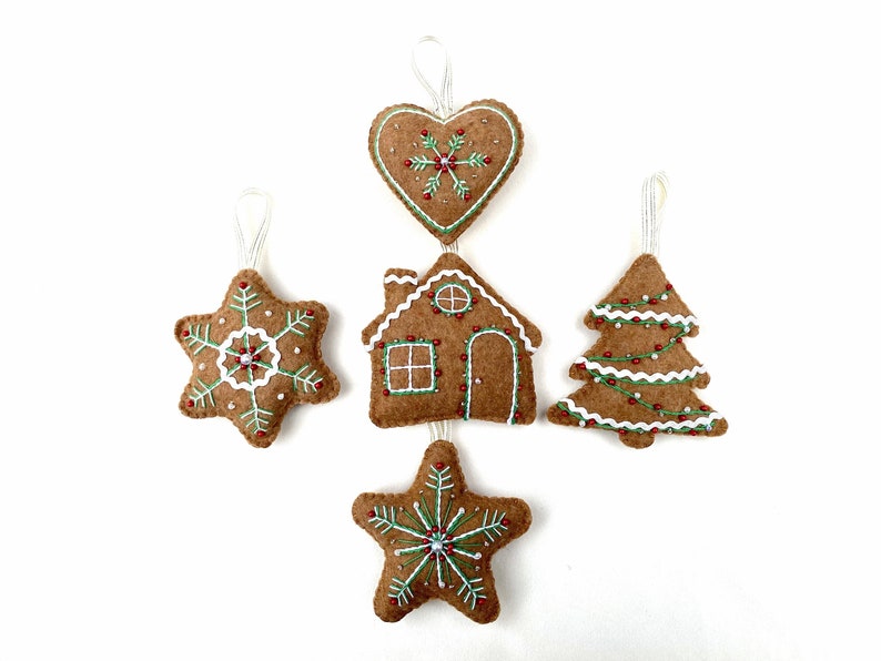 May include: Set of five felt gingerbread ornaments, including a heart, a house, a star, a tree, and a snowflake. Each ornament is decorated with white icing, green sprinkles, and red beads.