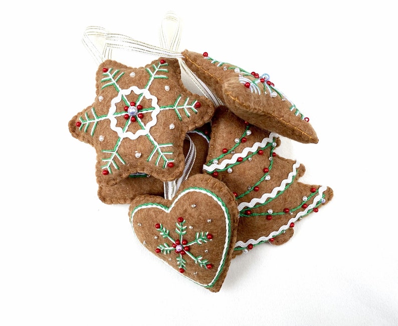 May include: Five felt Christmas ornaments shaped like a star, heart, and Christmas tree. The ornaments are brown with green and white embroidery and red and white beads.