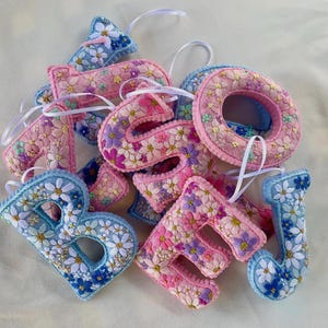 May include: A collection of plush, three-dimensional fabric letters featuring embroidered floral patterns in shades of pink, blue, and white. Each letter is finished with a white ribbon loop for hanging, showcasing intricate stitching of daisies.