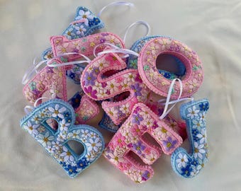 May include: A collection of plush, three-dimensional fabric letters featuring embroidered floral patterns in shades of pink, blue, and white. Each letter is finished with a white ribbon loop for hanging, showcasing intricate stitching of daisies.