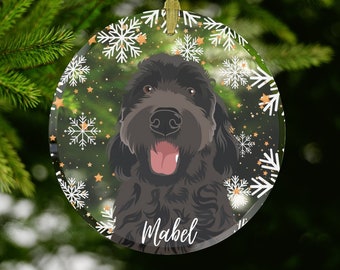 May include: A circular Christmas decoration with a black dog portrait, tongue extended, framed by white snowflakes and gold stars. The name "Mabel" is in white script. A gold ribbon is attached for hanging.