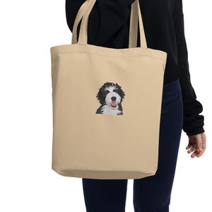 Op de afbeelding: Beige draagtas met een geborduurd beeld van een zwart-witte hond. De hond steekt zijn tong uit. De tas heeft twee lange handvatten en is gemaakt van een stevige stof. Het ontwerp is eenvoudig en opvallend.