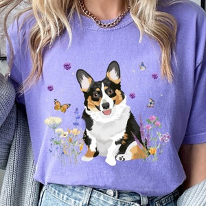 Może przedstawiać: Fioletowy T-shirt z grafiką przedstawiającą psa rasy corgi siedzącego na polu kwiatów i motyli. Pies jest czarny, biały i brązowy.