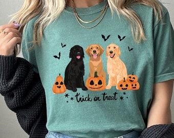 May include: A light green t-shirt featuring a graphic of three dogs and pumpkins. The dogs are a black labrador, a golden retriever, and a blonde golden retriever. The text on the shirt reads "Trick or Treat".