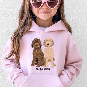 Może przedstawiać: Różowa bluza z kapturem z grafiką dwóch psów, jednego brązowego i jednego jasnobrązowego, siedzących obok siebie. Tekst "LACEY & QUINN" jest nadrukowany pod psami.
