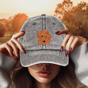 May include: A gray denim baseball cap with a brown embroidered doodle of a dog's head on the front.