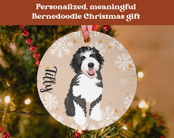 May include: A round, cream-colored Christmas ornament with a black and white Bernedoodle dog illustration. The ornament has a white snowflake pattern and gold stars. The dog's name, "Tilly", is written in black cursive on the ornament.
