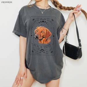 Pode incluir: Camiseta cinza escuro com um retrato de cachorro marrom em uma moldura decorativa. O texto "SOCK THIEF" está abaixo do cachorro. A camiseta tem mangas curtas e um corte folgado.