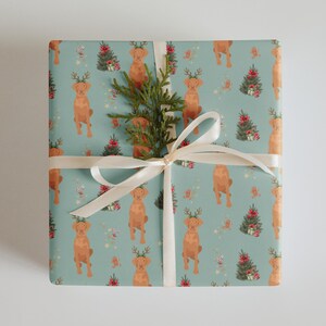 Könnte beinhalten: Verpacktes Geschenk mit hellblauem Geschenkpapier mit einem sich wiederholenden Muster aus braunen Hunden mit Geweih, Weihnachtsbäumen und Geschenken. Ein weißes Band und ein Zweig Grün schmücken die Oberseite. Ideal zum Schenken an Feiertagen.