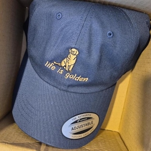 May include: Navy blue adjustable baseball cap with a gold embroidered image of a dog and the phrase "life is golden." The cap has a curved brim and a metal adjustable buckle.