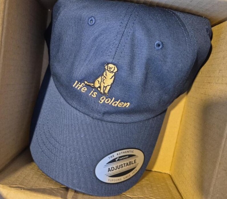 May include: Navy blue adjustable baseball cap with a gold embroidered image of a dog and the phrase "life is golden." The cap has a curved brim and a metal adjustable buckle.