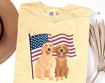 May include: A pale yellow Comfort Colors t-shirt with a graphic of two Golden Retrievers and an American flag. The shirt has a crew neck and short sleeves. A delicate star pendant necklace is draped across the shirt.