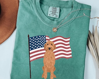 May include: Light green t-shirt featuring a graphic of a golden doodle dog and an American flag. The shirt has a crew neck and short sleeves. The brand label "Comfort Colors" is visible on the shirt.