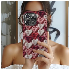 May include: A phone case featuring a repeating pattern of knitted hearts in shades of red, pink, and white. The case has a black camera module and is held in front of a person's face.