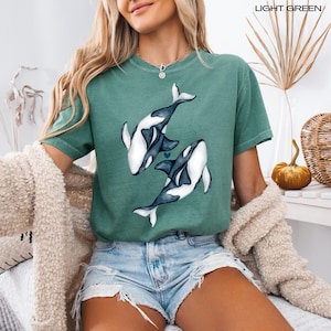 May include: A light green t-shirt featuring a graphic of two orcas swimming in a heart shape. The orcas are black and white.