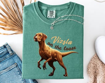 May include: A light green t-shirt featuring a brown Vizsla dog illustration in a dinosaur pose. The text "Vizsla on the loose" is printed in brown and gold on the shirt.