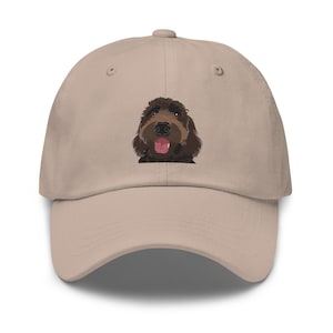 May include: A tan baseball cap featuring an embroidered image of a brown dog's face. The dog has a pink tongue and is smiling. The cap has a curved brim and a button on top.