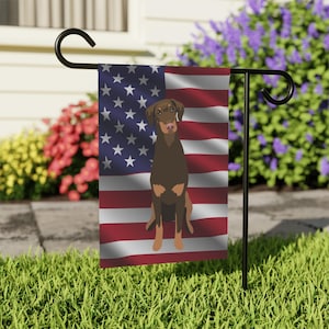 Peut inclure: Un drapeau de jardin avec le drapeau américain et une illustration d'un Doberman Pinscher brun. Le drapeau est affiché sur un support en métal noir dans un jardin herbeux, avec des fleurs colorées en arrière-plan.