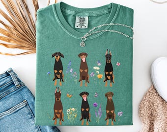 Può includere: Una maglietta verde chiaro con una grafica di sei Doberman Pinscher in piedi in un campo di fiori. La maglietta ha un'etichetta Comfort Colors nell'angolo in basso a sinistra.