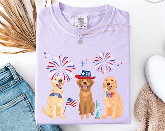 May include: Purple t-shirt featuring a design of three golden retrievers wearing patriotic hats and bow ties. The dogs are surrounded by fireworks and a small American flag. The shirt has the text "COMFORT COLORS" on the tag.