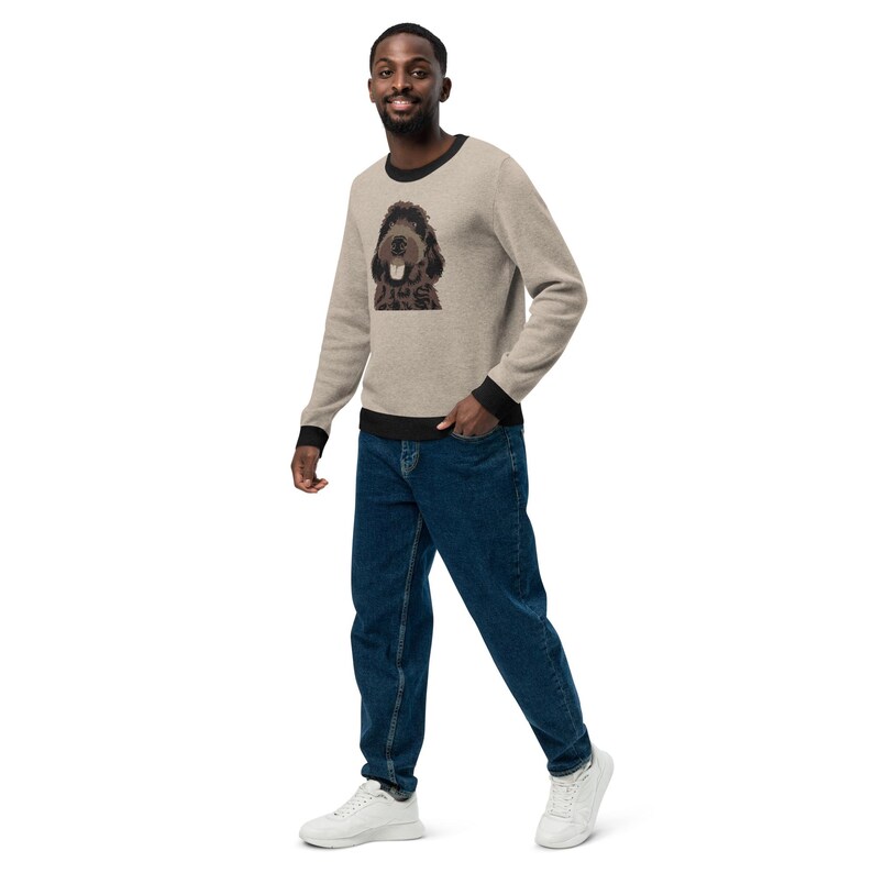 May include: A man wearing a beige sweater with a black trim and a brown dog illustration on the front. He is wearing blue jeans and white sneakers.