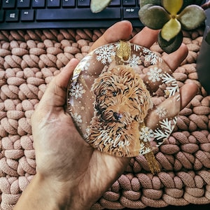 Pode incluir: Um enfeite de Natal de vidro transparente com uma fita dourada e um cão castanho e branco dentro. O nome do cão é "Milan" impresso no enfeite.