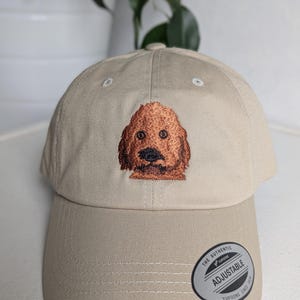 May include: A beige baseball cap with an embroidered brown dog on the front. The cap has an adjustable strap with a silver tag that reads "The Authentic Yupoong Adjustable Yupoong Since 1974".