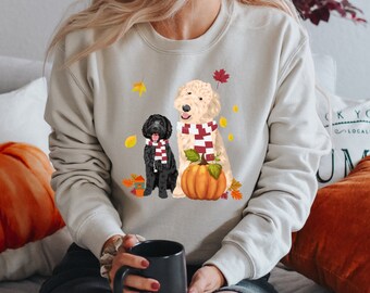 Könnte beinhalten: Ein hellgraues Sweatshirt mit einem Aufdruck von zwei Hunden, die Schals tragen und einen Kürbis. Die Hunde sind ein schwarzer Pudel und ein cremefarbener Pudel. Der Aufdruck ist von Herbstblättern umgeben.