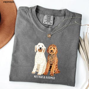 May include: A grey Comfort Colors t-shirt with a graphic of two dogs, one white and one brown, and the text "ARTHUR & BOOMER" underneath. The shirt has a crew neck and short sleeves. A silver necklace with a pendant is draped on the shirt.