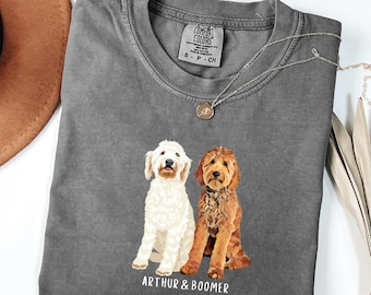 May include: A gray Comfort Colors t-shirt featuring a graphic of two dogs, one white and one brown, with the text "ARTHUR & BOOMER" below. The shirt has a crew neck and short sleeves. A silver necklace with a pendant is draped on the shirt.