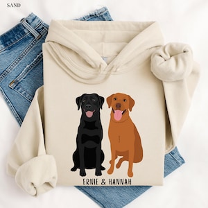 Peut inclure: Un sweat à capuche couleur crème avec un labrador noir et un labrador brun assis côte à côte. Le texte "ERNIE & HANNAH" est imprimé sous les chiens.