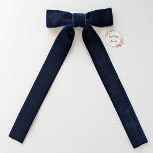 May include: A navy blue velvet bow with long, trailing tails. The bow is centrally positioned against a white backdrop. The text "Ruthies Bows" is within a floral wreath on the right.