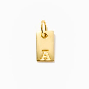 May include: A gold-coloured rectangular pendant featuring the letter "A". The pendant has a small loop at the top for attaching to a chain or bracelet. The background is plain white.
