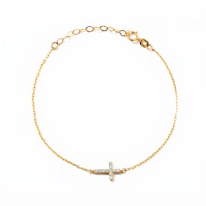 May include: A delicate gold chain bracelet featuring a small, horizontal cross pendant centered on the chain. The cross is embellished with tiny, clear, sparkling stones set along its surface. The bracelet includes a secure lobster claw clasp.