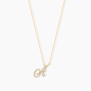 May include: A gold-colored necklace with a cursive letter "A" pendant. The pendant and chain are gold, and the letter is encrusted with small, sparkling stones. The necklace hangs against a white background.