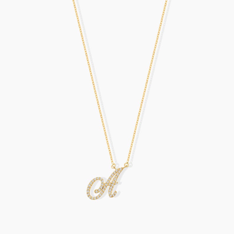 May include: A gold-colored necklace with a cursive letter "A" pendant. The pendant and chain are gold, and the letter is encrusted with small, sparkling stones. The necklace hangs against a white background.