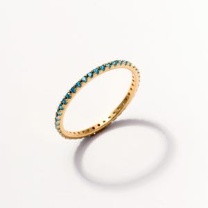 May include: A thin gold band ring featuring a continuous row of small, round, bright blue gemstones set along the outer circumference. The metal has a polished finish, and the stones are evenly spaced, creating a delicate, circular jewelry piece.