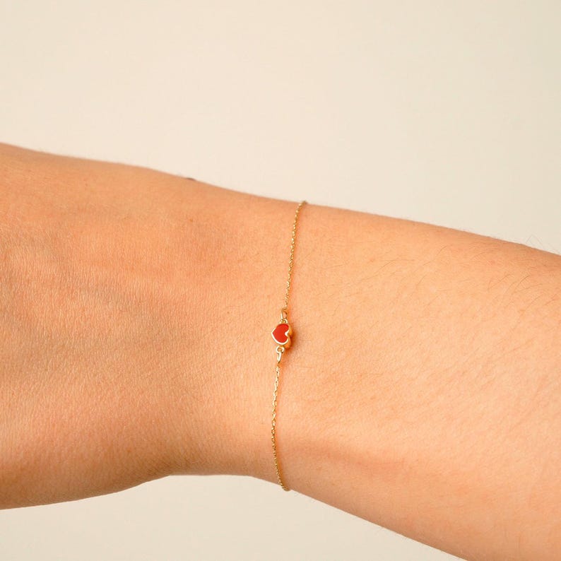 May include: A delicate gold bracelet featuring a small, red heart charm. The charm is centered on a thin gold chain. The bracelet is shown on a person's wrist, against a neutral background. Jewelry.